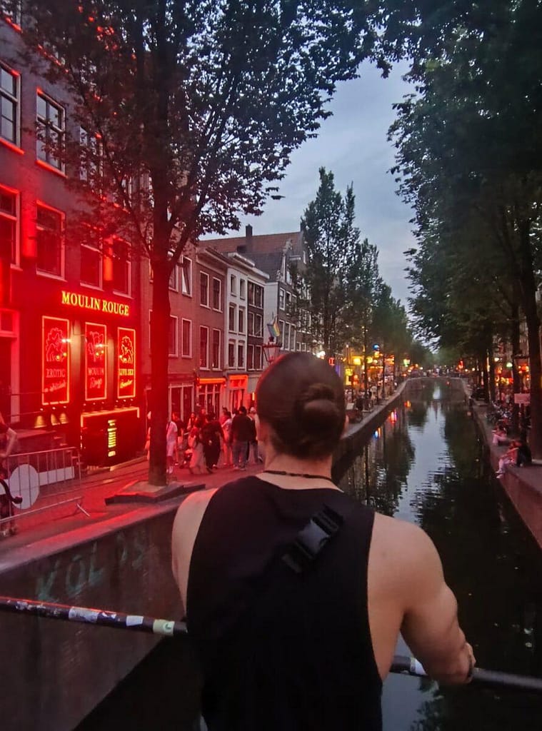 Mężczyzna patrzący na kanał w Amsterdamie wieczorem, z widocznym neonem Moulin Rouge w Dzielnicy Czerwonych Latarni.