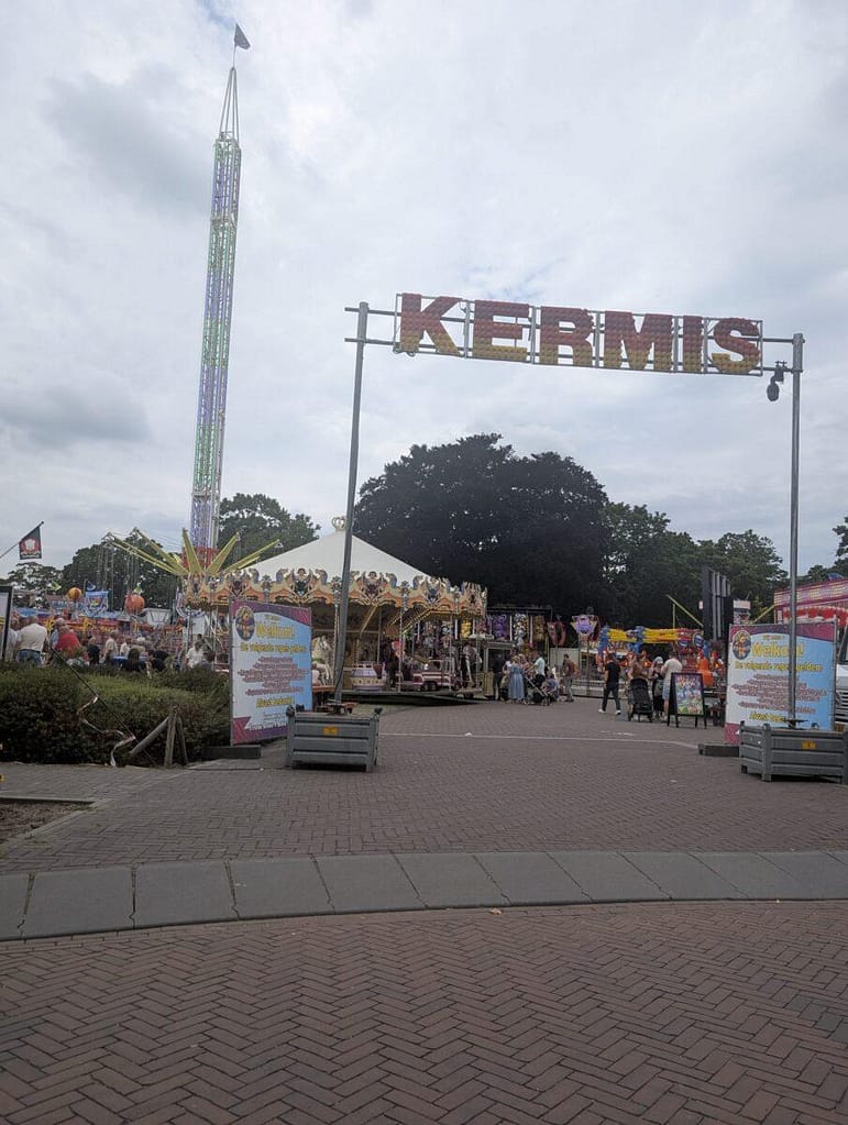 Widok na tradycyjne holenderskie wesołe miasteczko Kermis z dużą karuzelą i wysoką wieżą swobodnego spadku pod pochmurnym niebem.