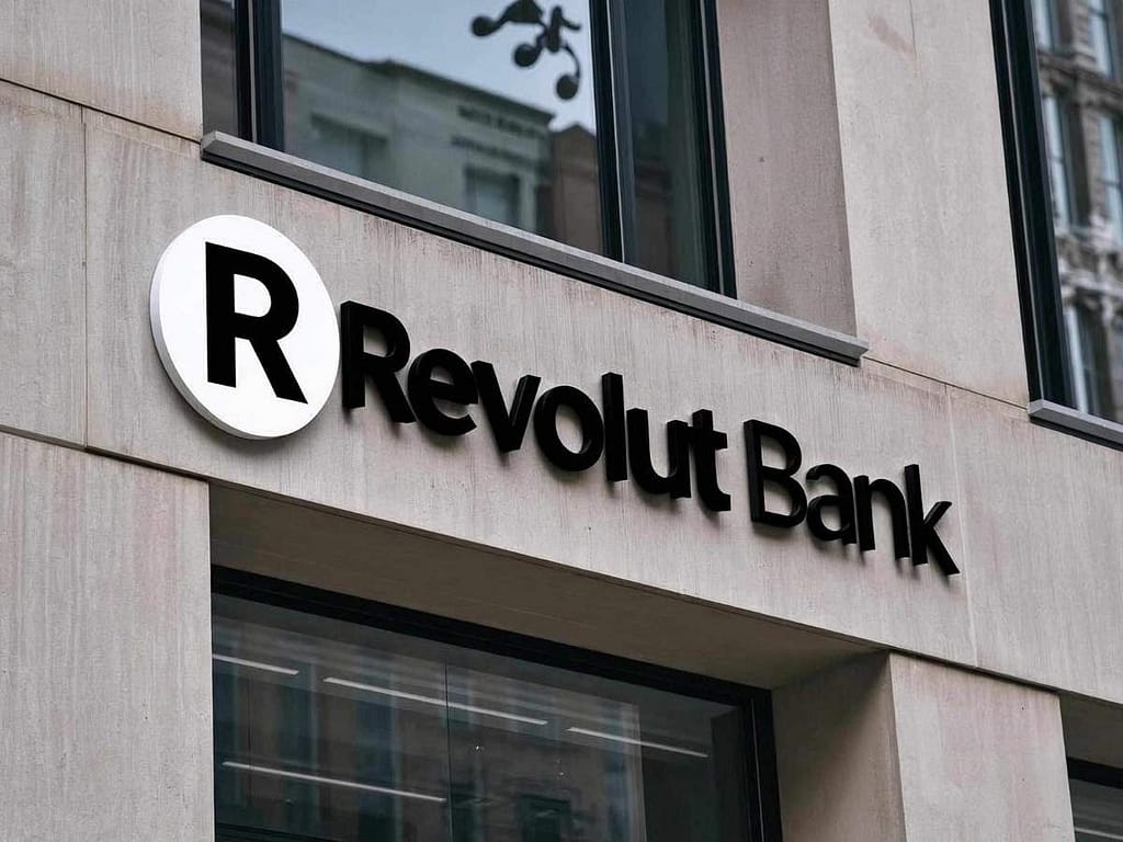 Czarny napis Revolut Bank z okrągłym logo na jasnej fasadzie nowoczesnego budynku biurowego.